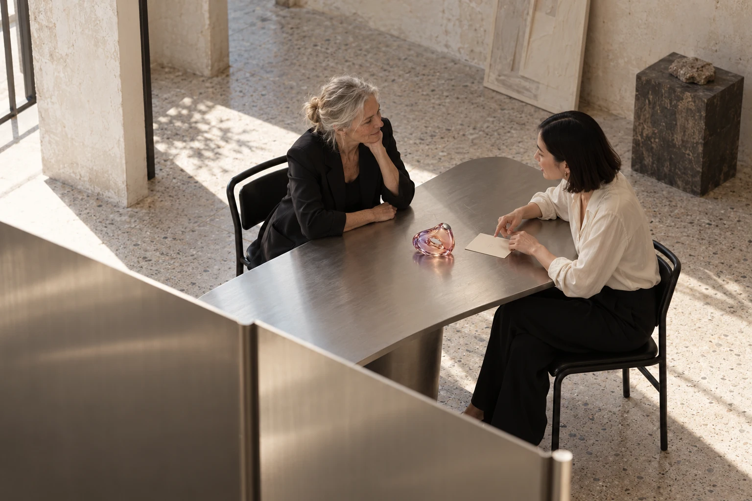 Two women in conversation around the SINGULÆ vessel in a light stone and brushed metal atelier